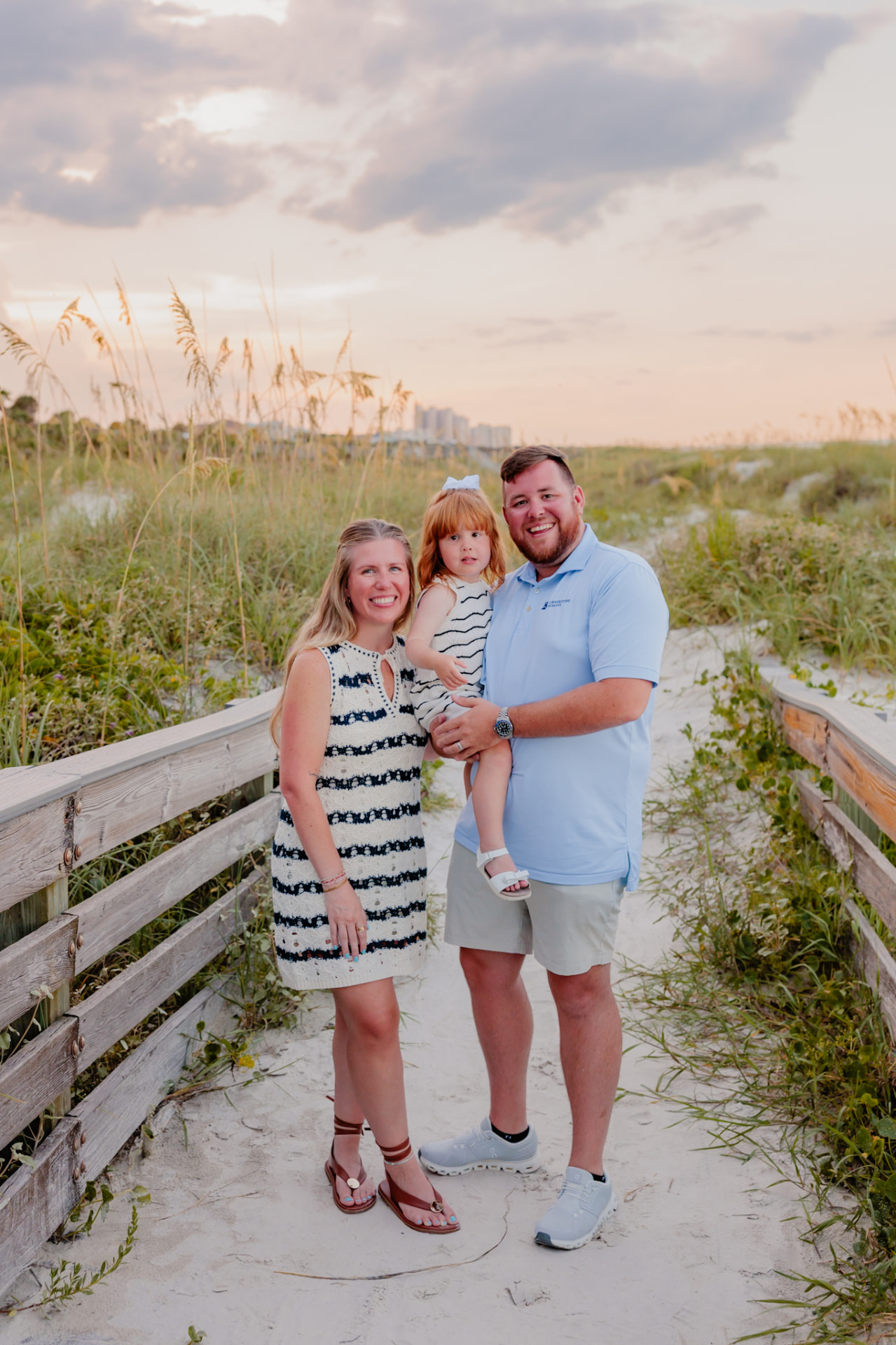 Trevor Seay holding his young daughter while walking with his wife down a sandy coastal boardwalk at sunset