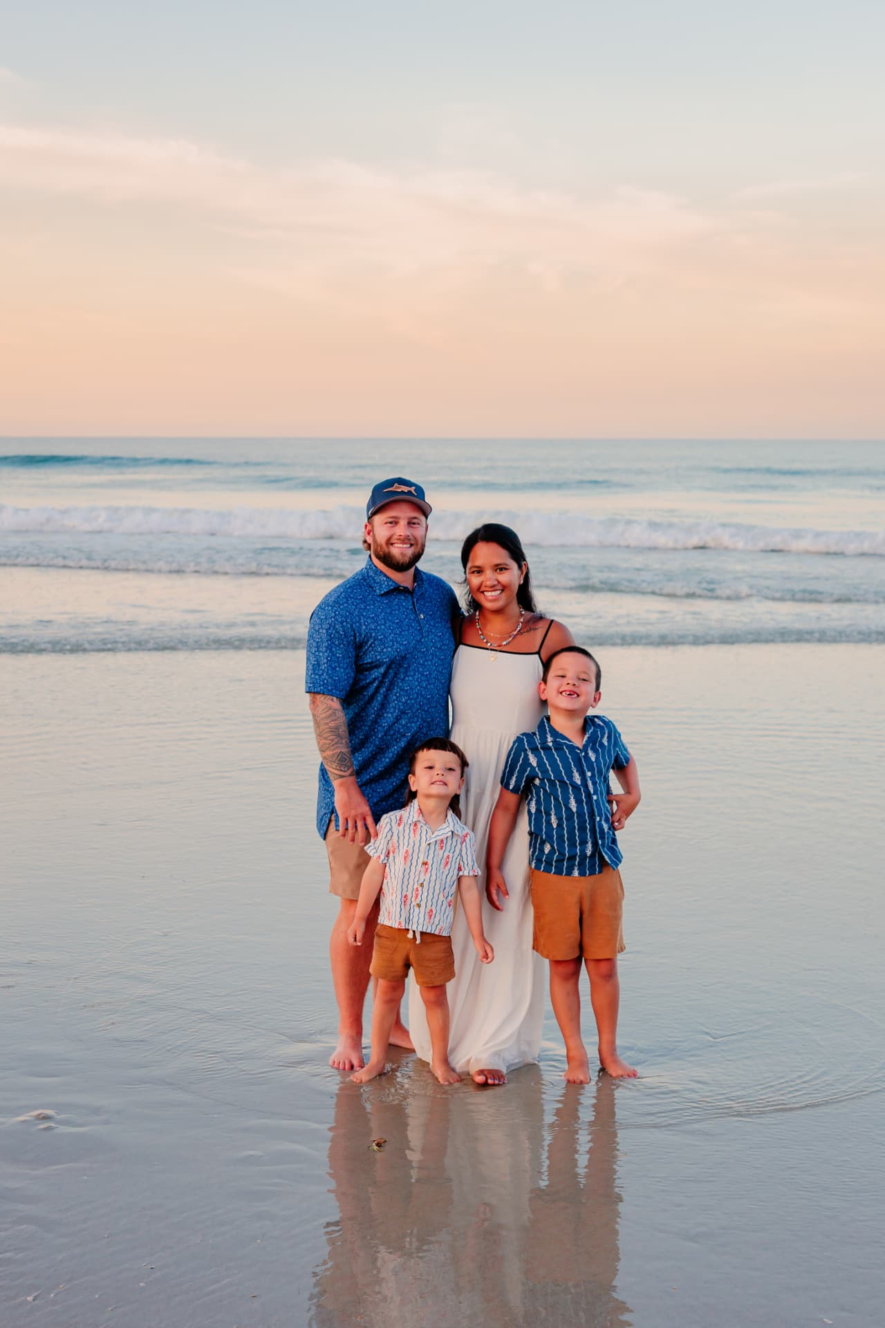 Garrett Cox with his wife and two young sons standing barefoot together in the shallow surf on a beach at sunset